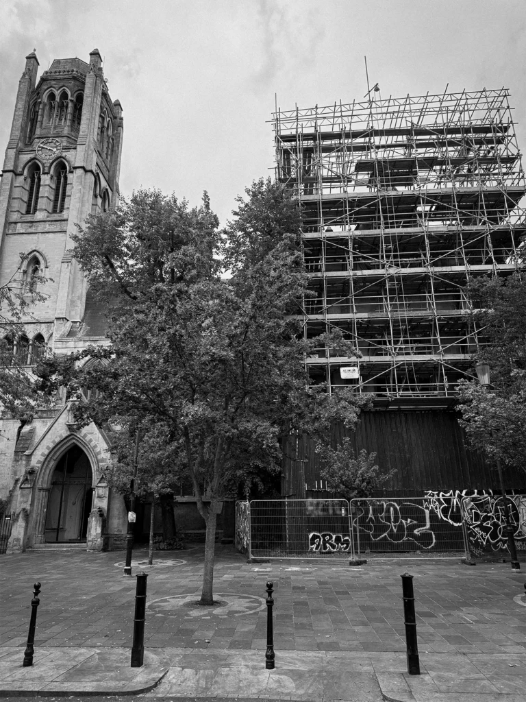 Black and white photo of All Saints Church in Notting Hill, London, with its Gothic clock tower on the left and the nave wrapped in scaffolding on the right, a tree in front and graffiti-covered hoardings at street level.