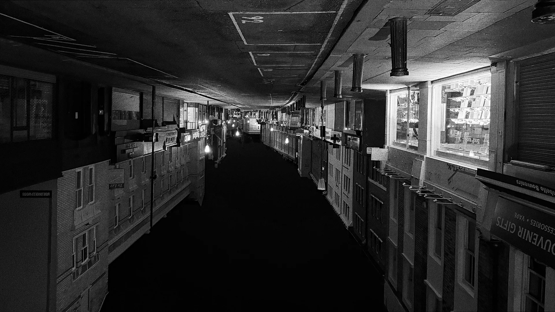 Black-and-white photo of an empty high street at night, turned on its head, one shop window glowing among shuttered shopfronts.