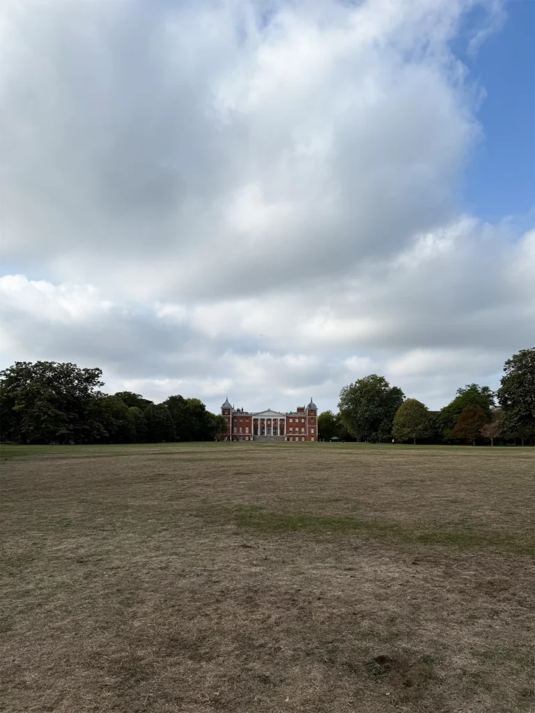 A wide, sun-dried lawn stretches towards Osterley House, a red-brick mansion with a white columned portico and corner turrets, framed by trees beneath a vast sky of billowing white clouds with patches of blue.